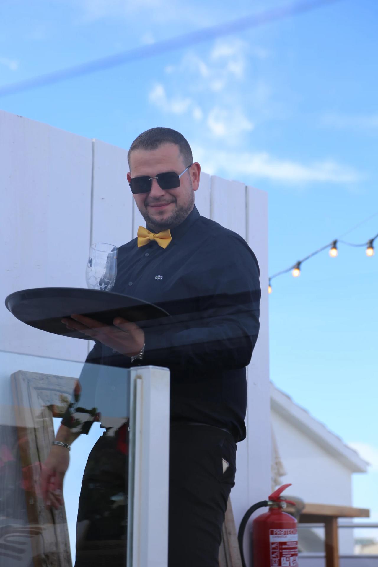 Waiter holding a tray with a glass, wearing sunglasses and a bow tie.