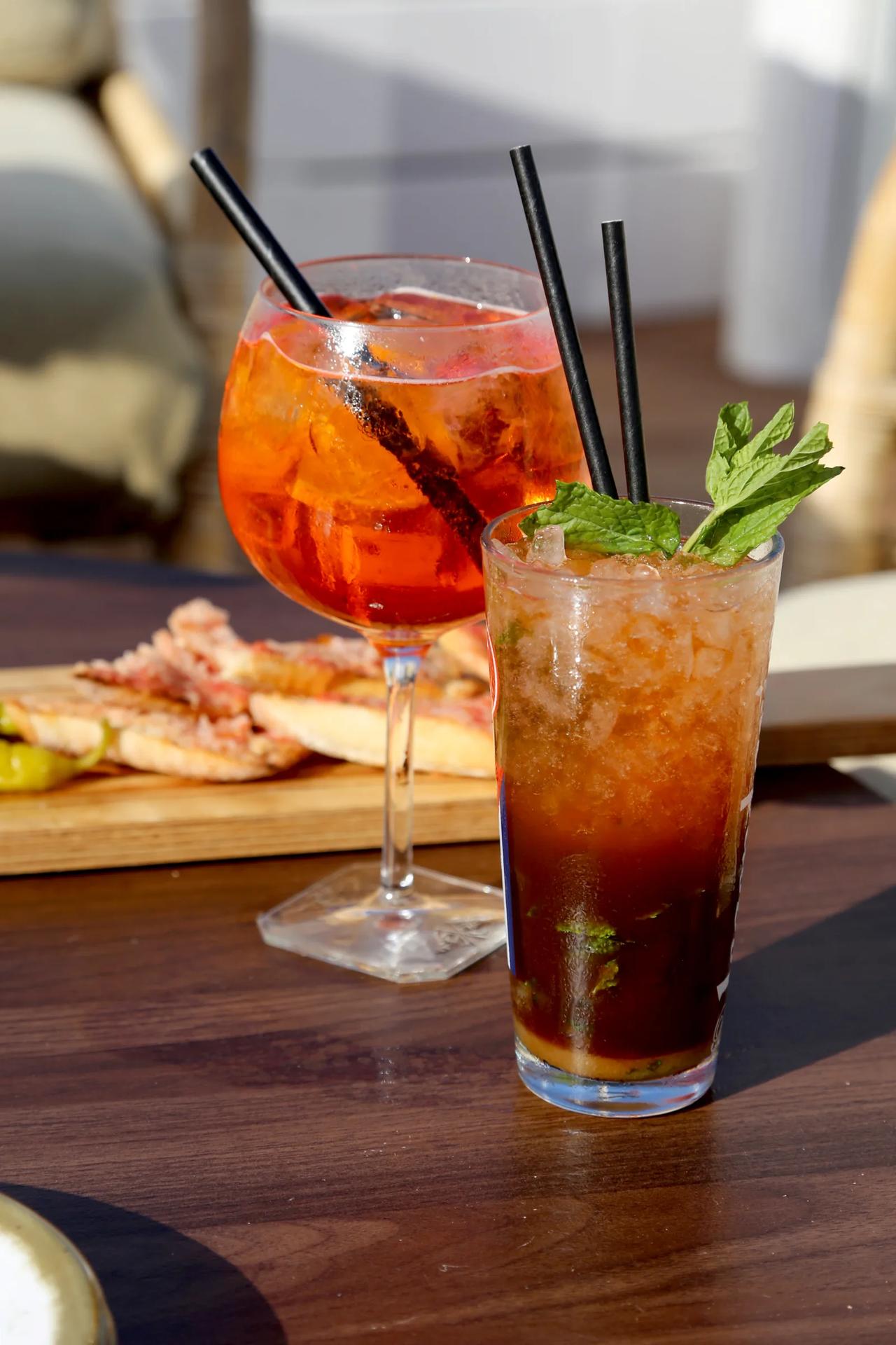 Two cocktails on a wooden table with snacks in the background.