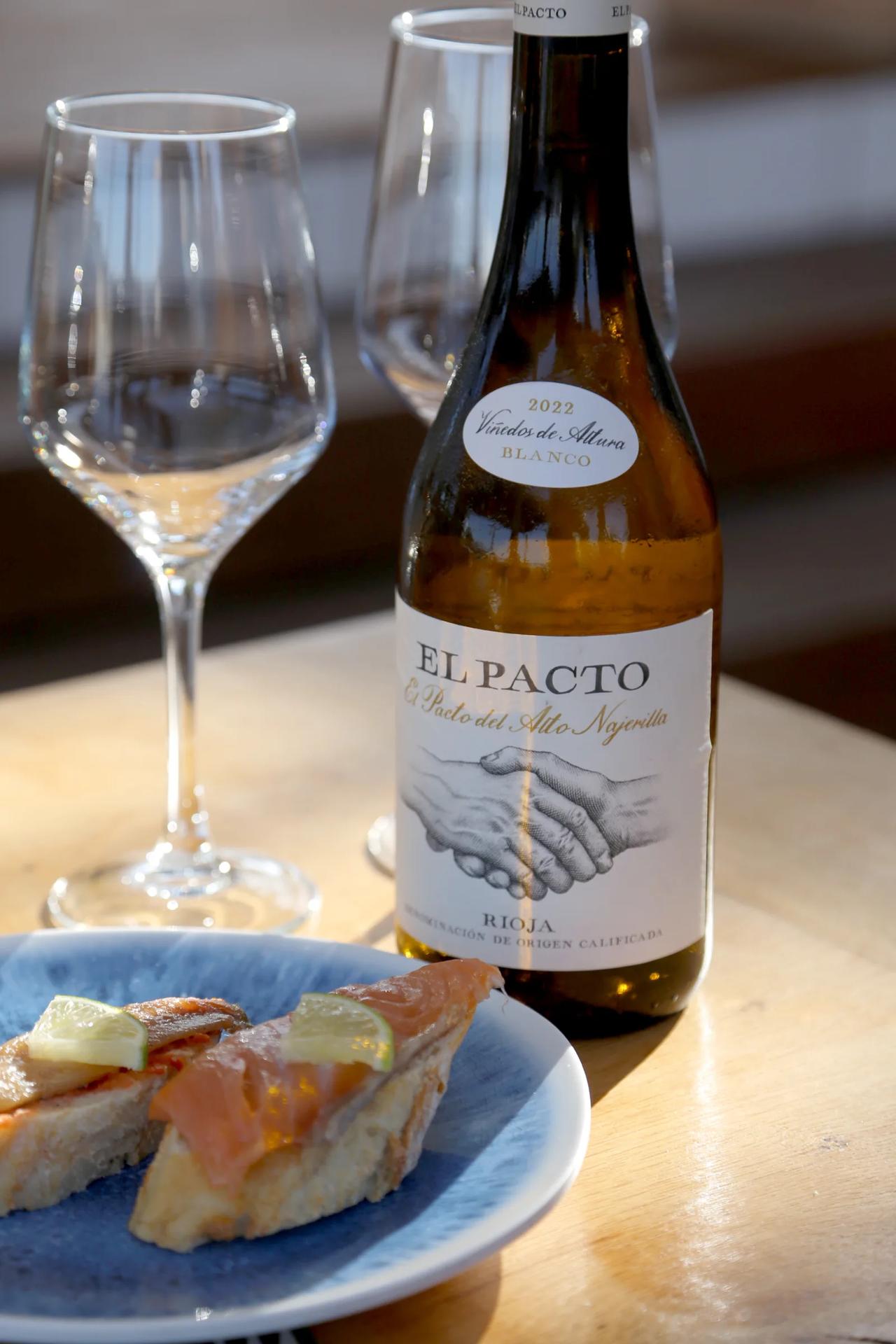 A bottle of El Pacto wine with glasses and a plate of food.