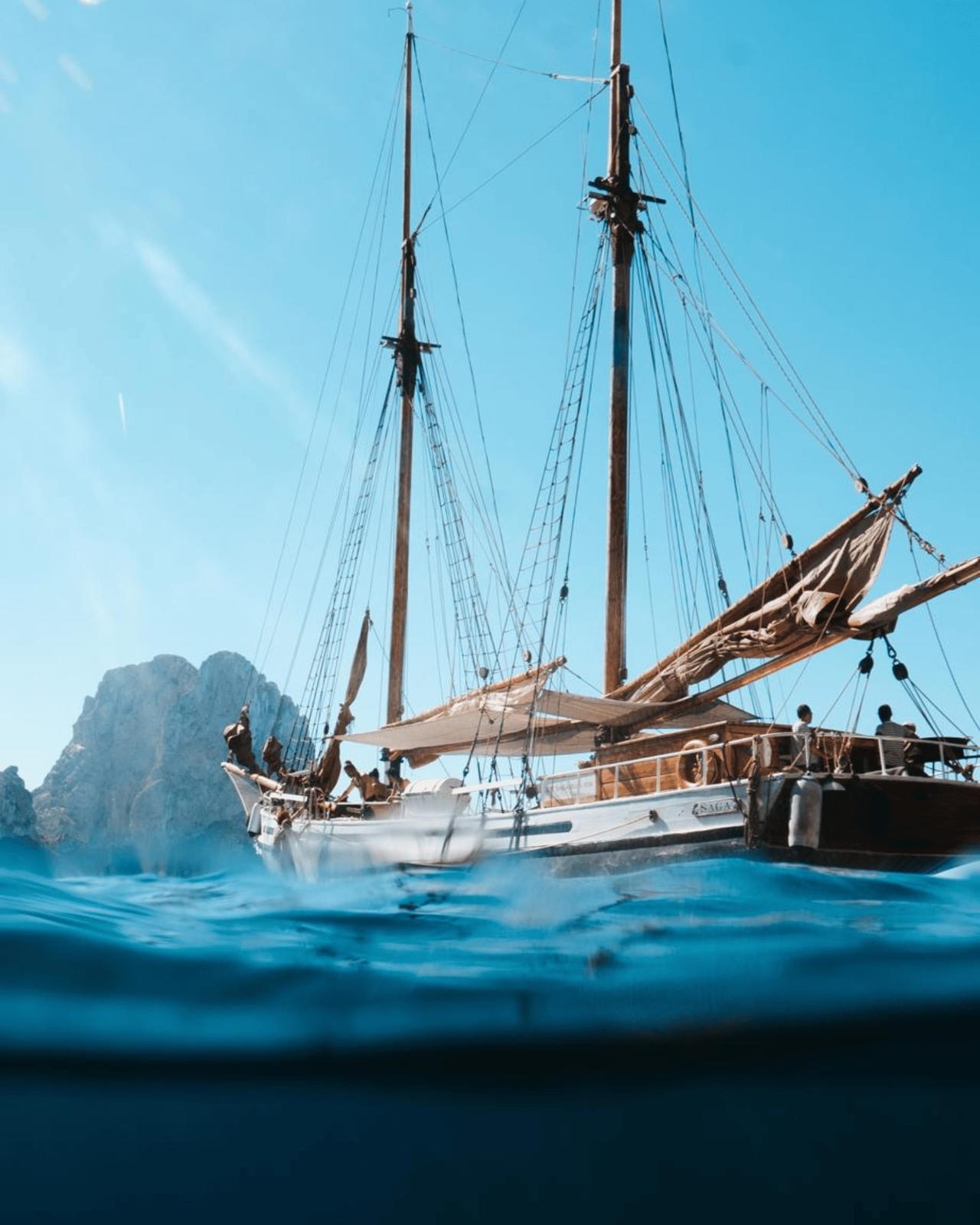 Sailing boat with tall masts near rocky coastline under clear blue sky.