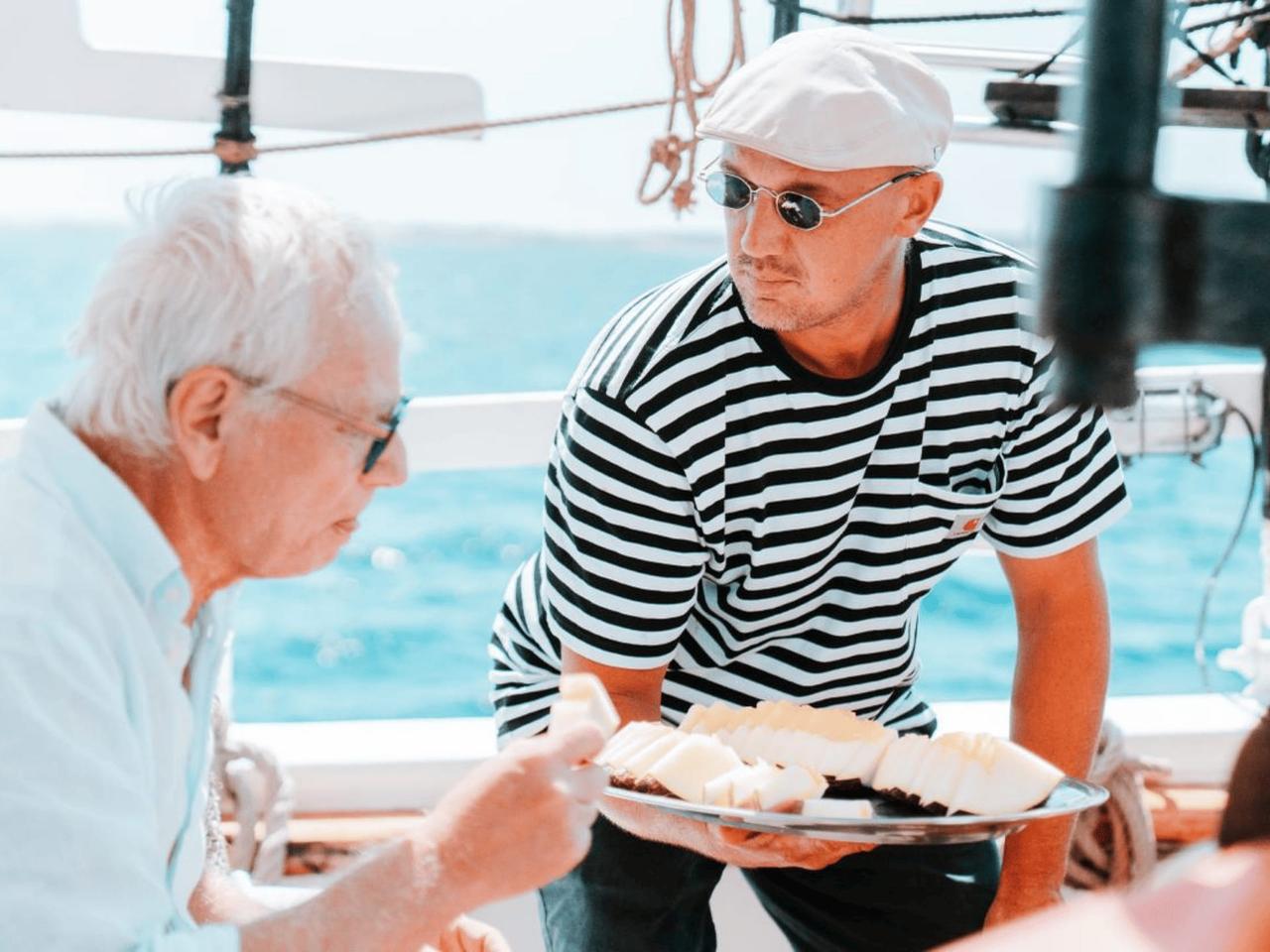 Two men on a boat enjoying food, one serving a platter of fruit.