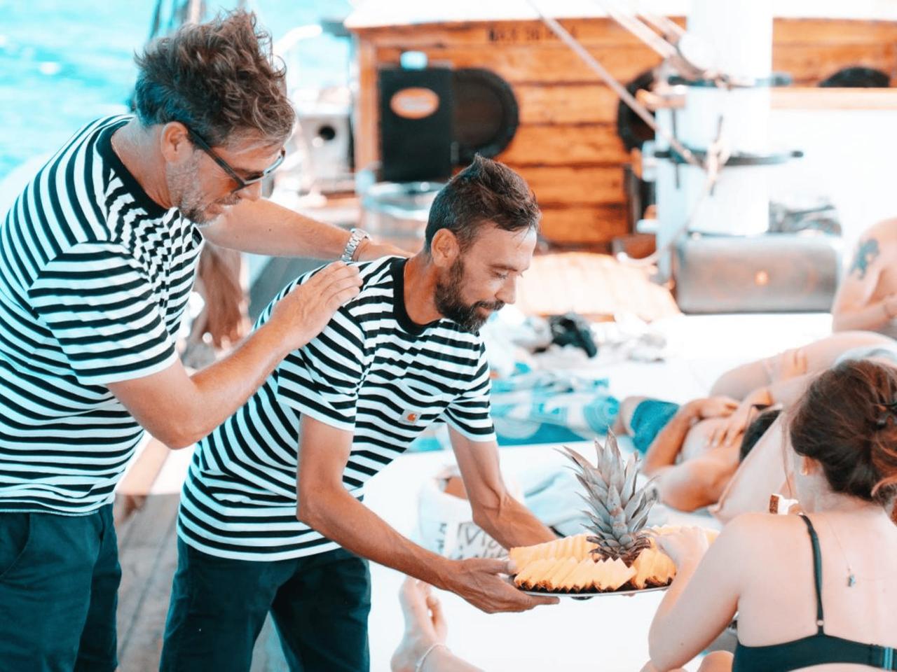 Two men in striped shirts serving a pineapple on a boat.