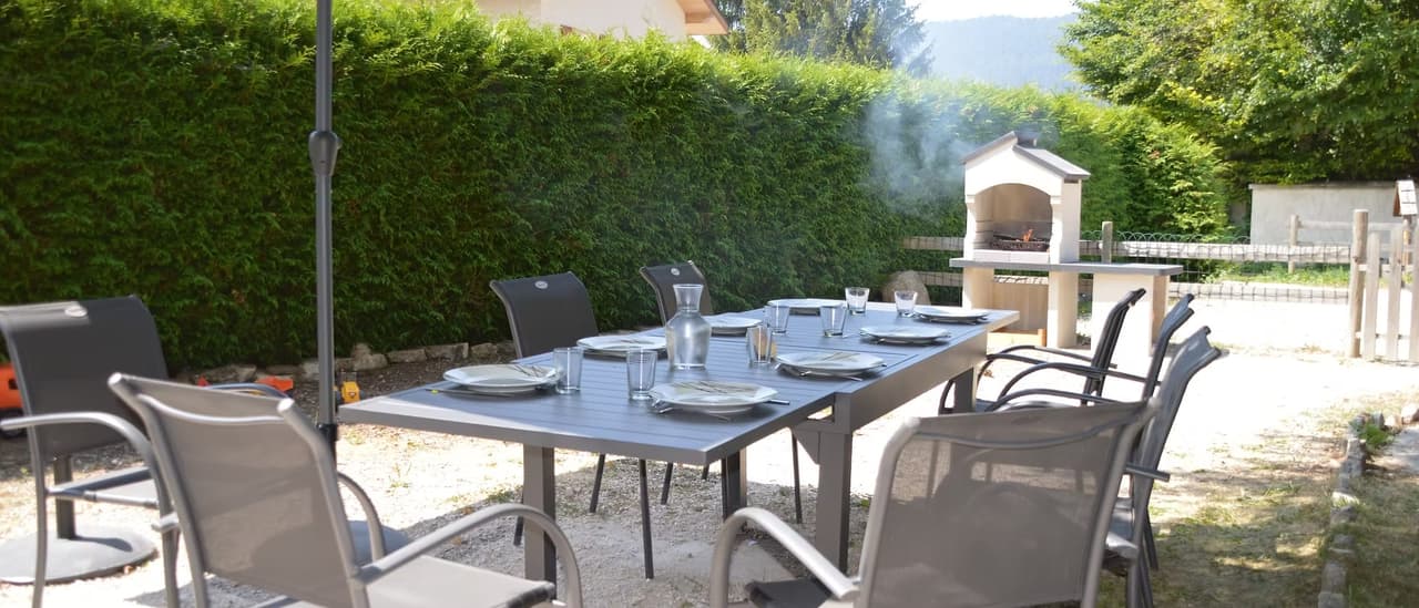 Table de jardin avec chaises, barbecue et verdure en arrière-plan.